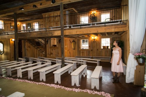 A young girl in a pink dress stands in a rustic wooden venue with benches and floral decorations, preparing for an event.