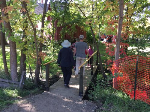 Two people walk across a wooden bridge surrounded by trees with colorful autumn leaves, leading to a red building.