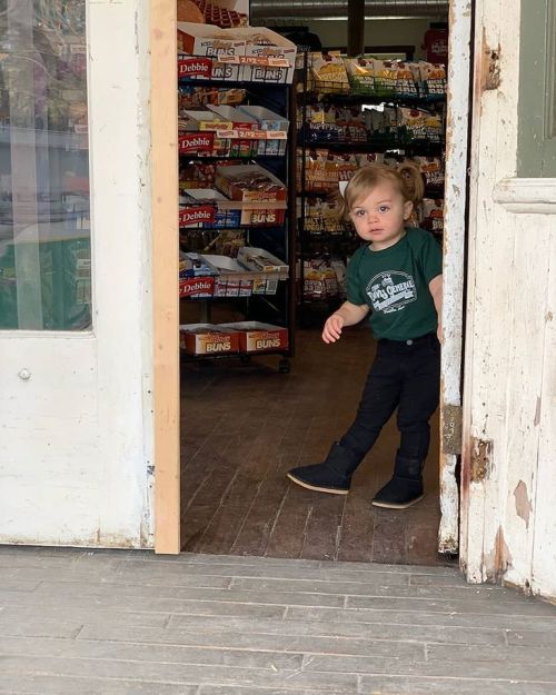 A young child in a green shirt and black pants stands at the entrance of a store, surrounded by shelves of snacks.