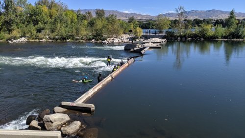 A group of people in wetsuits near a river, navigating a small rapid with trees and mountains in the background.