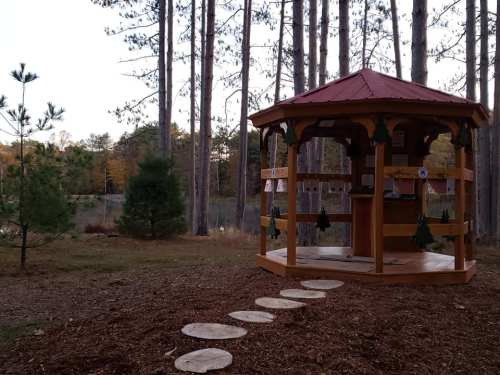 A wooden gazebo with a red roof surrounded by trees and a stone path, near a calm body of water.