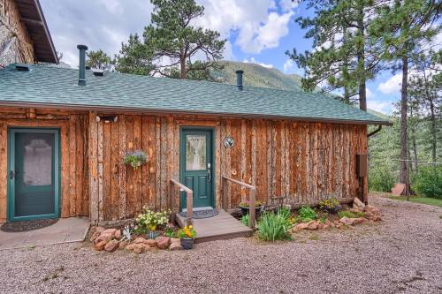A rustic wooden cabin with green doors, surrounded by trees and mountains, featuring a small porch and flower beds.