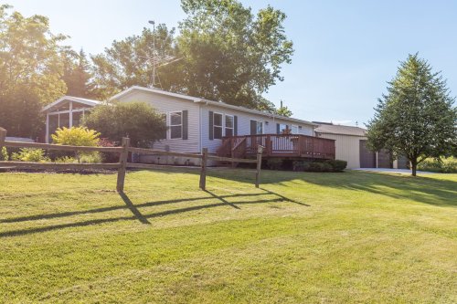 A single-story house with a deck, surrounded by green grass and trees, with a garage in the background.