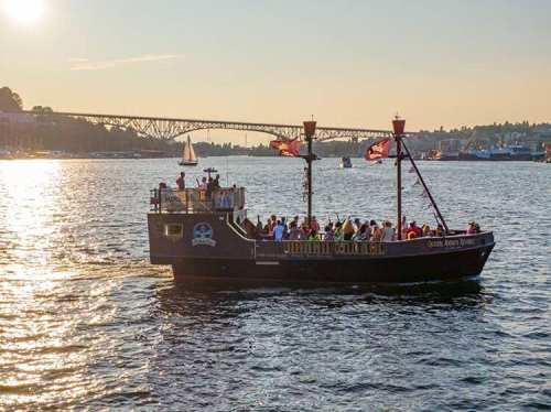 A pirate-themed boat with passengers sails on a shimmering waterway, with a bridge in the background at sunset.