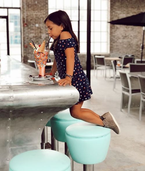 A young girl in a navy dress leans over a counter, admiring a colorful ice cream sundae in a bright, modern setting.