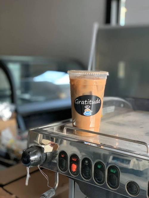 A clear cup of iced coffee with a "Gratitude" label sits on a counter near a coffee machine.