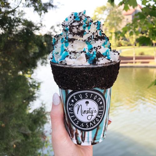 A hand holds a colorful dessert cup topped with whipped cream, crushed cookies, and blue sprinkles by a scenic water view.