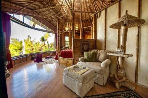 Cozy interior of a bamboo hut with a bed, armchair, and large windows showcasing a lush outdoor view.