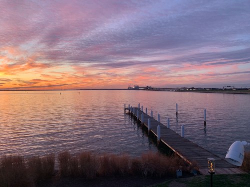 A serene sunset over calm waters, with a wooden pier extending into the horizon and colorful clouds in the sky.