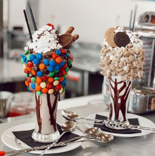 Two colorful ice cream sundaes topped with whipped cream, candies, and cookies, served in tall glasses with spoons.