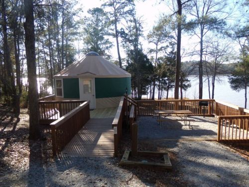 A yurt with a wooden deck near a lake, surrounded by trees and a picnic area.