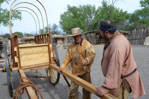 Two men in traditional attire discuss a wooden wagon in a historical outdoor setting with trees in the background.