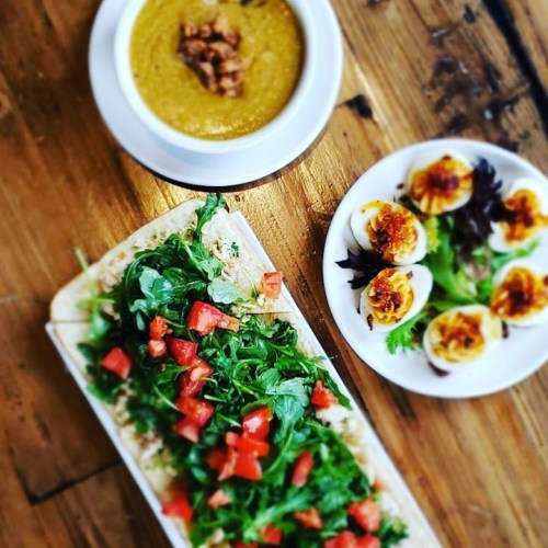 A wooden table with a bowl of soup, a plate of deviled eggs, and a flatbread topped with greens and diced tomatoes.
