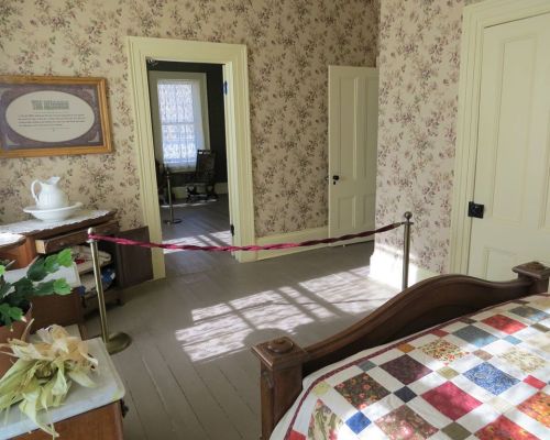 A vintage bedroom with floral wallpaper, a quilted bed, and a doorway leading to another room, separated by a rope barrier.