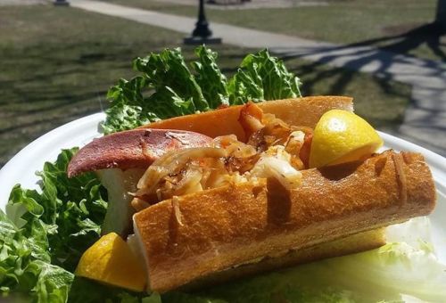 A lobster roll on a bed of lettuce, garnished with lemon wedges, served on a white plate outdoors.