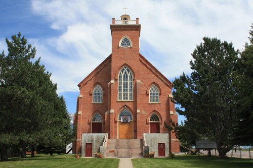 A red brick church with a tall steeple, large windows, and a grassy area surrounded by trees under a blue sky.