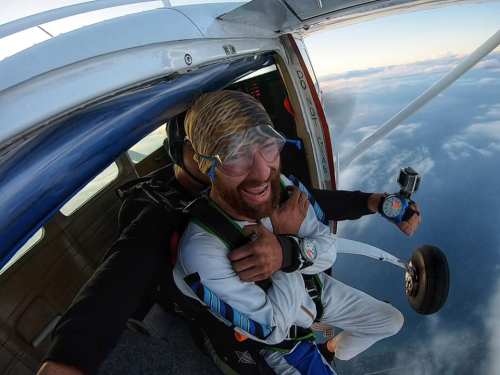 A person in skydiving gear smiles while preparing to jump from an airplane, with clouds visible below.