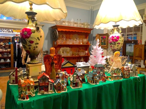 A display of decorative houses and figurines on a green tablecloth, with lamps and shelves of collectibles in the background.