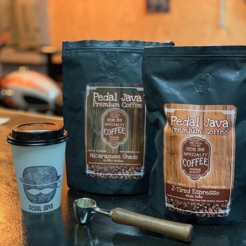 Two bags of Pedal Java coffee, a coffee cup with a mustache logo, and a coffee scoop on a wooden surface.