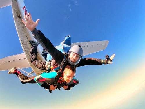 A tandem skydiver leaps from a plane, arms outstretched against a clear blue sky.