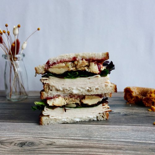 A stacked sandwich with layers of turkey, lettuce, and sauce, next to a small piece of bread and a vase of dried flowers.