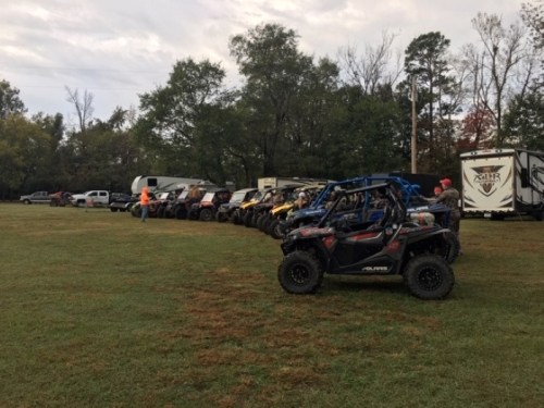 A row of parked ATVs and trucks on a grassy field, with trees in the background and a cloudy sky above.