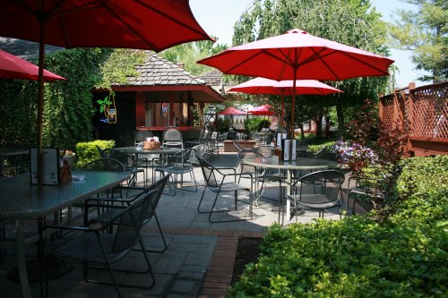 Outdoor dining area with tables and red umbrellas, surrounded by greenery and a cozy atmosphere.