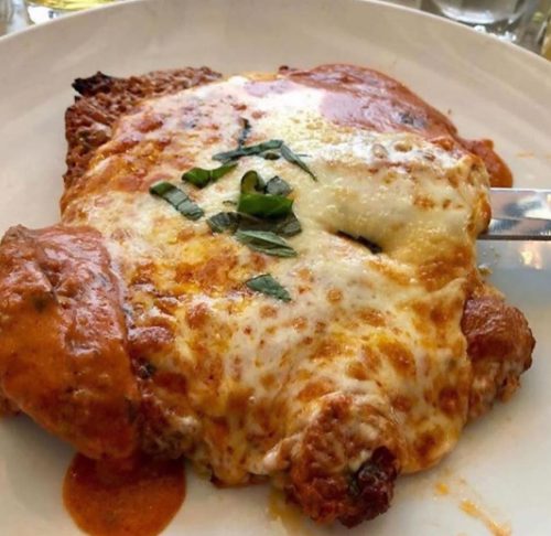 A close-up of a cheesy chicken dish topped with marinara sauce and fresh basil on a white plate.