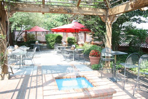 A sunny outdoor patio with tables, red umbrellas, and a small fountain surrounded by greenery.
