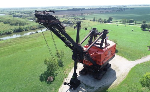 A large orange excavator stands on a grassy area near a river, surrounded by fields and trees under a clear sky.