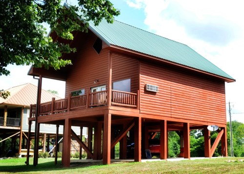 A wooden house on stilts with a green metal roof, surrounded by trees and grass under a blue sky.