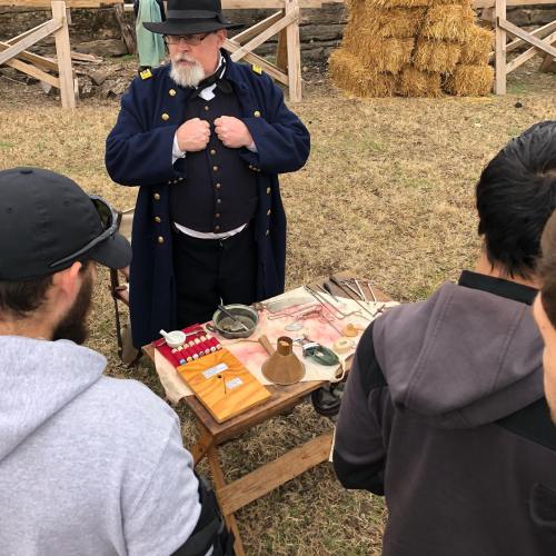 A historical reenactor in a blue coat demonstrates tools to an audience at an outdoor event. Hay bales are in the background.