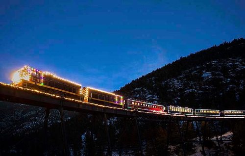 A brightly lit train decorated with holiday lights travels across a bridge at dusk, surrounded by mountains.