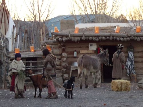 Two people interact with a goat and a donkey outside a rustic wooden structure, with lanterns glowing in the background.