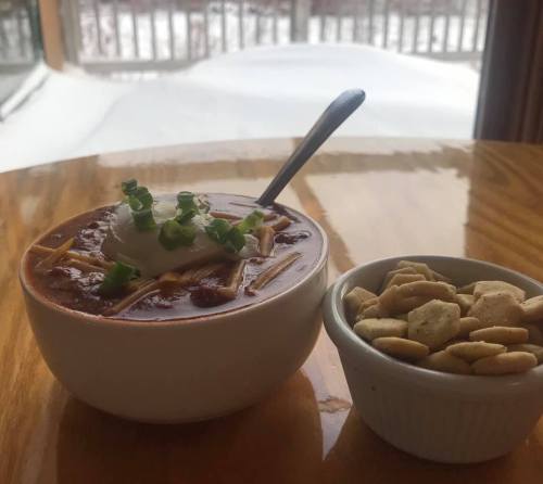 A bowl of chili topped with sour cream and green onions, alongside a small dish of crackers, with snow visible outside.