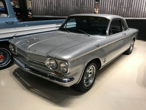 A classic silver coupe car with a sleek design, parked indoors against a wooden backdrop.