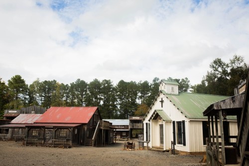 A rustic western town with wooden buildings, a church with a cross, and a cloudy sky in the background.