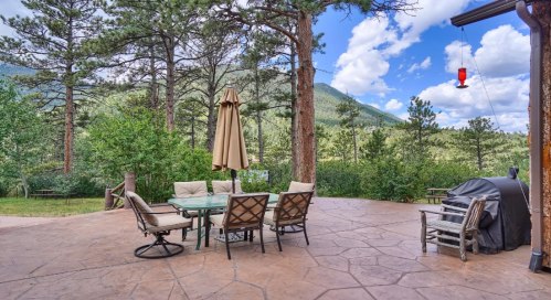 A patio with a glass table and chairs, an umbrella, and a grill, surrounded by trees and mountains under a blue sky.