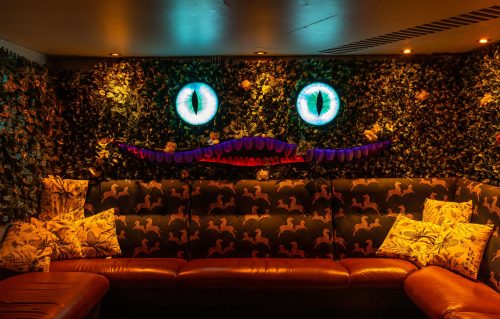 A cozy lounge with a green wall of leaves, featuring large blue eyes and a whimsical mustache above a brown sofa.