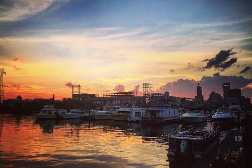 Sunset over a harbor filled with boats, reflecting vibrant colors on the water, with city skyline in the background.