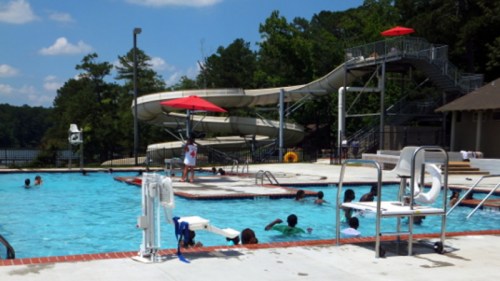 A sunny pool scene with children swimming, a water slide, and lifeguard chair surrounded by trees.
