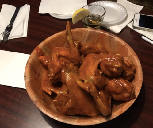 A wooden bowl filled with saucy chicken wings, accompanied by a lemon wedge and a glass of whiskey on the side.