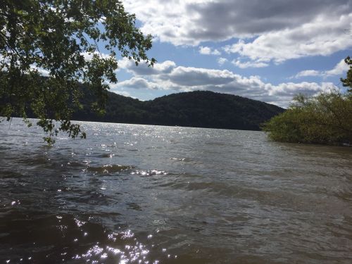 A serene river scene with sparkling water, green hills in the background, and a partly cloudy sky.