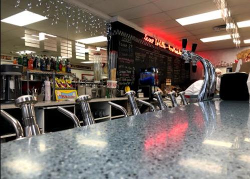 A diner counter with shiny taps, colorful drinks, and a menu board in the background, creating a nostalgic atmosphere.