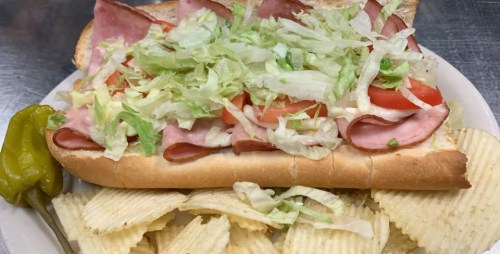 A sub sandwich loaded with ham, lettuce, and tomatoes, served with a side of potato chips and a pepper.
