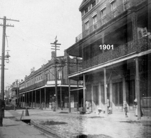 Historic street scene from 1901, featuring buildings, power lines, and pedestrians in a bustling urban setting.