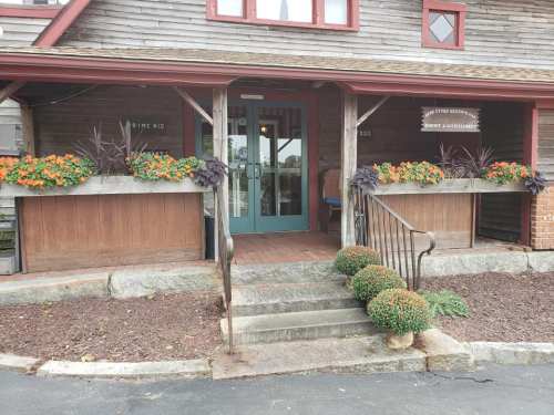 A rustic building entrance with green doors, flower planters, and a welcoming porch surrounded by colorful flowers.