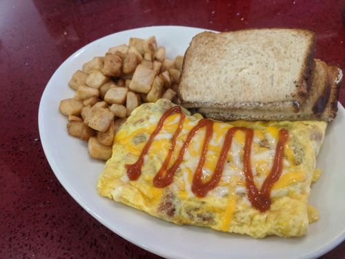 A plate with a cheesy omelet drizzled with ketchup, served with diced potatoes and two slices of toast.