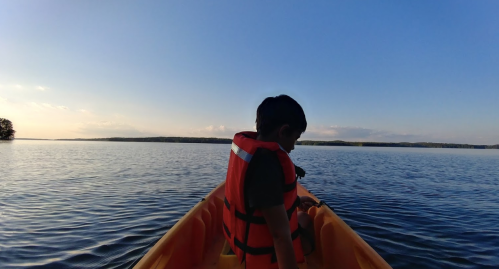A child in a life jacket sits in a kayak on a calm lake, with a clear blue sky and distant trees in the background.