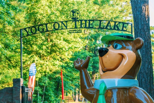 A cheerful bear statue waves under a sign reading "Yogi on the Lake," surrounded by lush green trees.